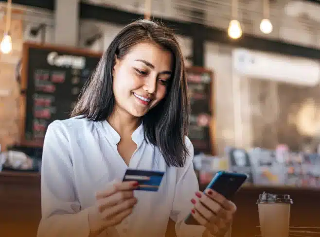 A happy female is making an online payment on her mobile through her card details while holding the card in hand.